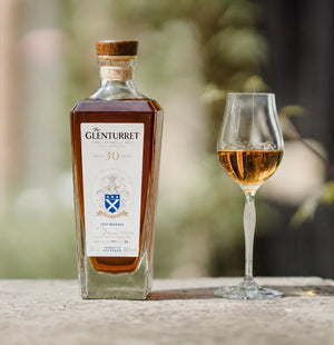 A bottle of The Glenturret 30 Years Single Malt Scotch whisky stands on a stone surface next to a stemmed glass filled with the golden amber liquid, set against a softly blurred outdoor background.