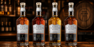 Four bottles of Finn Thomson whisky lined up on a wooden surface with a blurred bar background and a wooden barrel behind them.