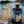 A dark amber bottle labeled FERAGAIA sits on a stone surface beside two tall glasses of a bubbly Ginger Ale & Feragaia with lime wedges, set against a blurred coastal background with water and rocks.