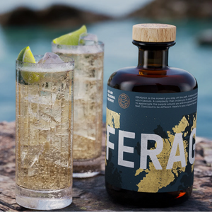 A dark amber bottle labeled FERAGAIA sits on a stone surface beside two tall glasses of a bubbly Ginger Ale & Feragaia with lime wedges, set against a blurred coastal background with water and rocks.