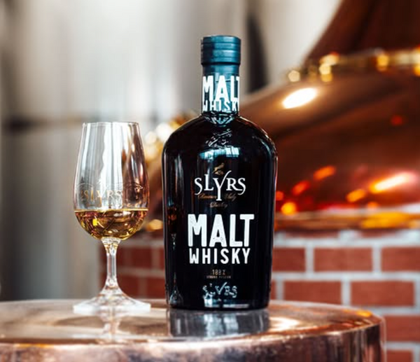 Bottle of Slyrs Malt Whisky with a glass on a wooden barrel against a blurred background